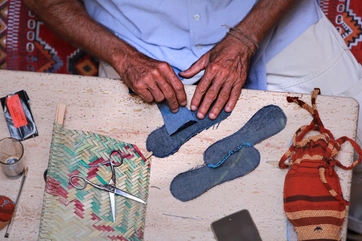 A maker's hands cutting denim insoles on a worn workboard — scissors, basket and an orange sac in frame.